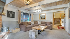 Un salon chaleureux avec un canapé d’angle, une table basse en bois et un élément décoratif en pierre. On peut voir une cheminée en briques et une grande fenêtre avec des rideaux.