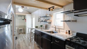 A modern kitchen with a built-in stove, a sink, and a countertop. There is a window with blinds, a ceiling fan, and a small stool. - The Merlot Bungalow Defiance Tiny Home on Trail (Defiance)