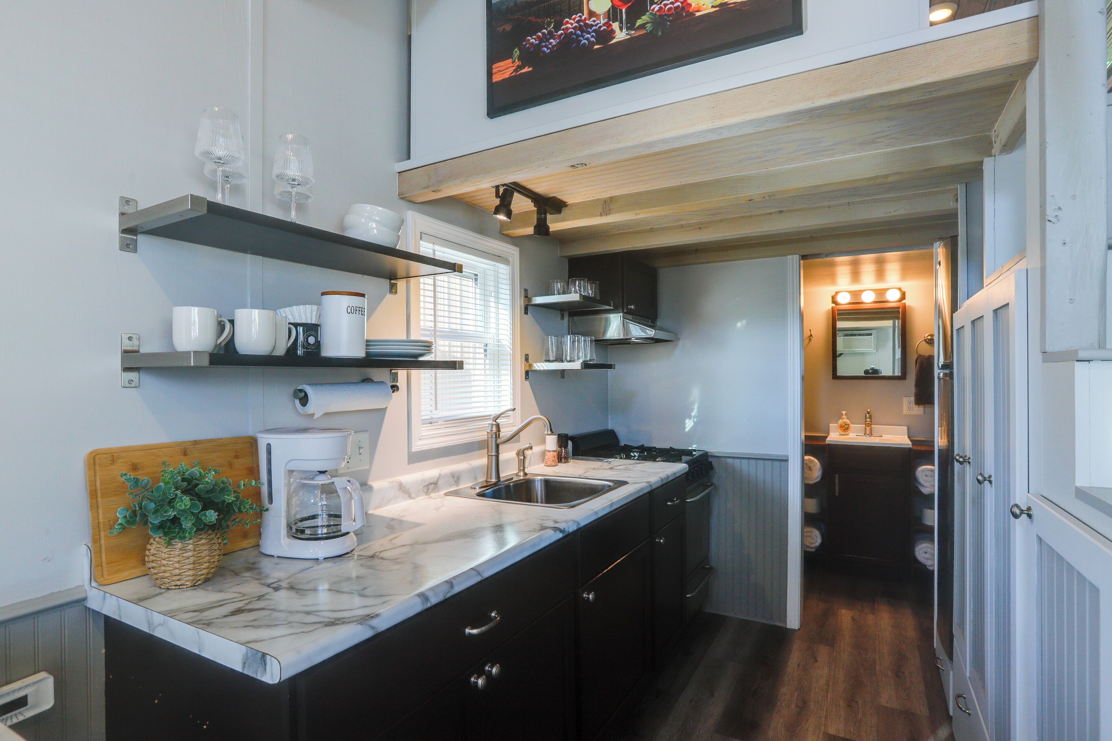 A compact kitchen with a coffee maker, shelves with glassware, and a sink.