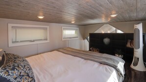 A bedroom with a wooden ceiling, two windows with blinds, a bed with a striped blanket, a bedside table, and a white fan. - The Merlot Bungalow Defiance Tiny Home on Trail (Defiance)