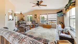 A cozy living room with a ceiling fan, a large window, a television, and a fireplace. - Lakefront Chiloquin Home w/ Scenic Deck! (Chiloquin)