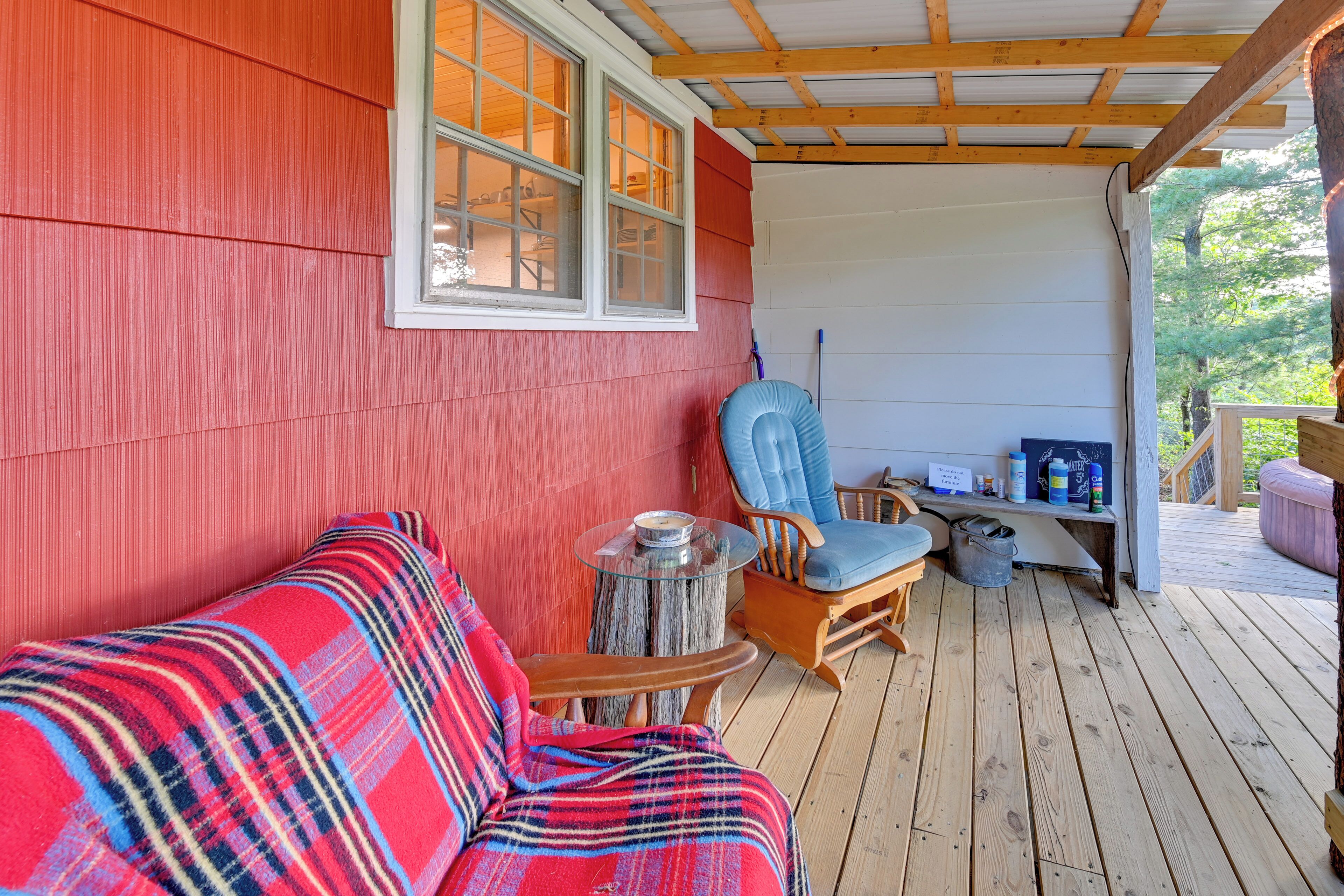 A covered porch with a red exterior, a wooden rocking chair, a blue cushioned chair, and a small table with a glass top.