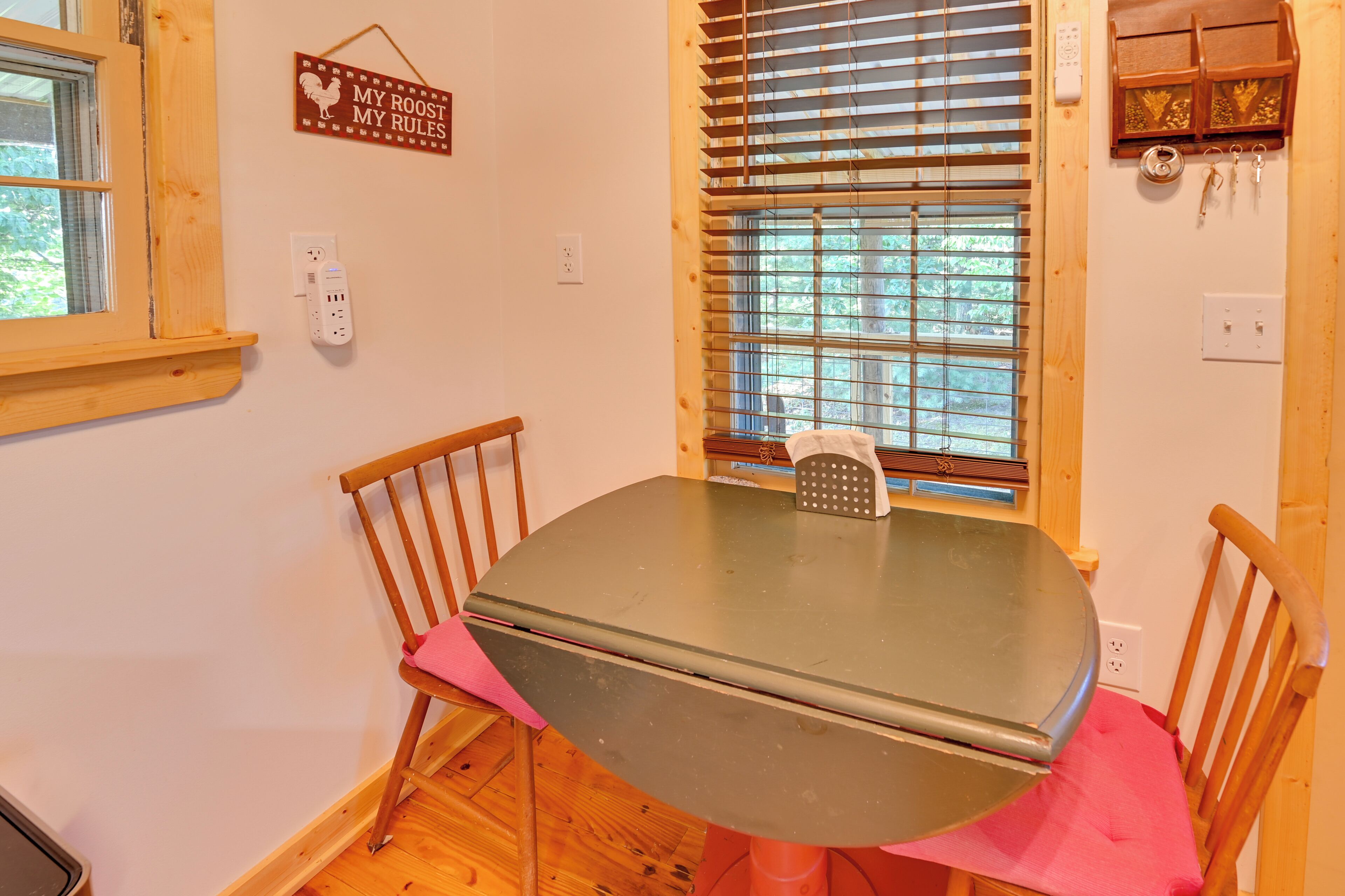 Un coin repas comprenant une table ronde, des chaises en bois, une fenêtre avec des stores et une prise électrique fixée au mur.