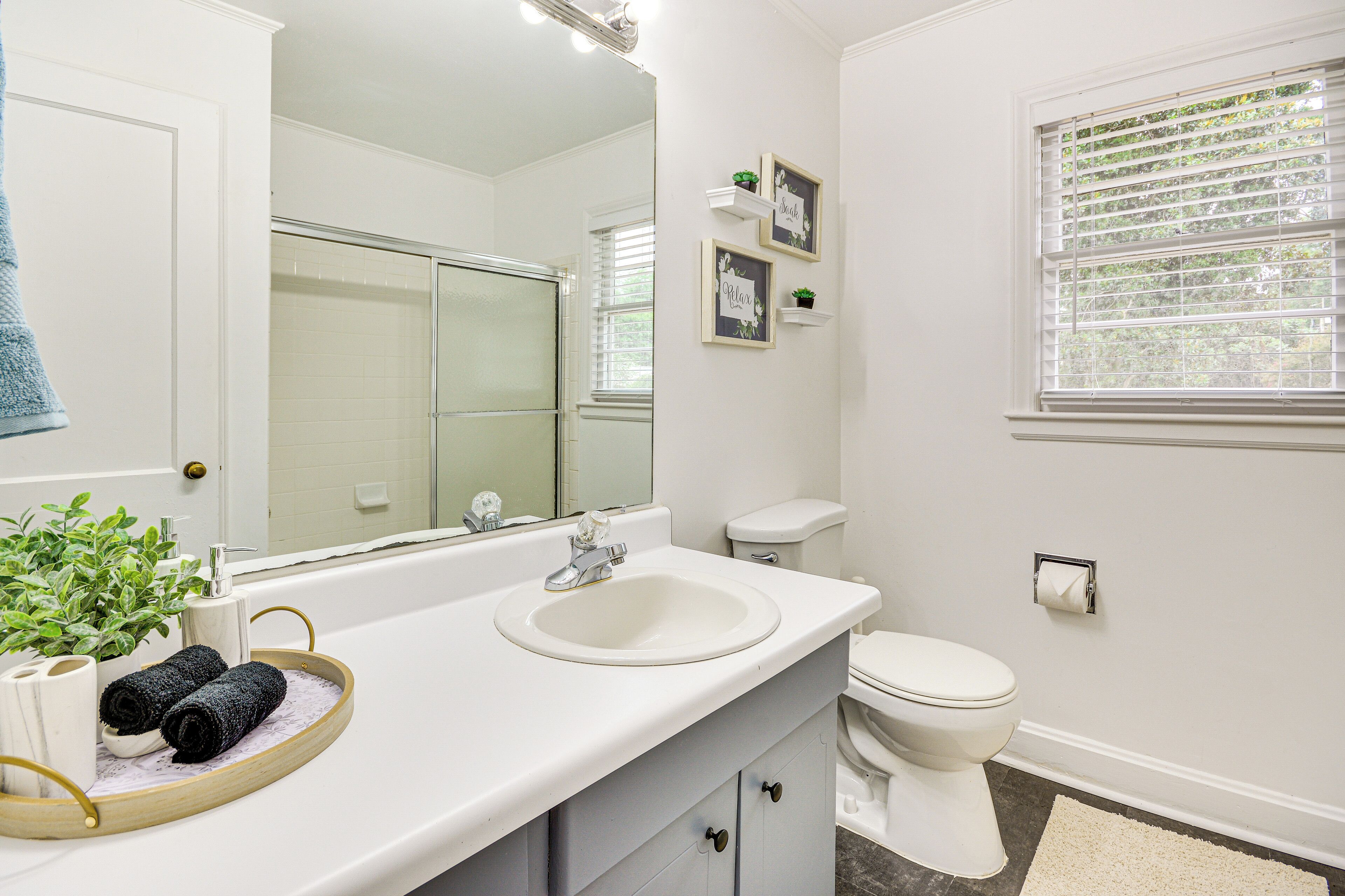 A bathroom with a white sink, mirror, and toilet. There is a towel rack with towels, a small plant, and a framed picture on the wall.