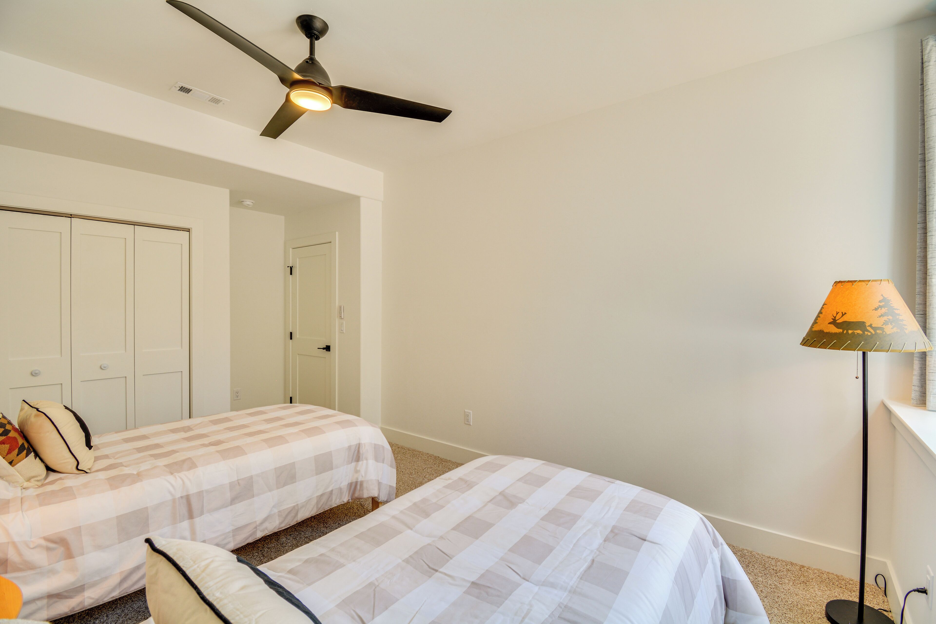 A bedroom with two beds, a ceiling fan, a lamp, and a door leading to another room.