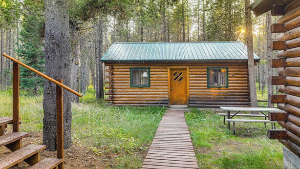 A log cabin with a wooden deck leading to a door, surrounded by trees. - Island Park Fishing Cabin - Walk to Henrys Fork! (Island Park)