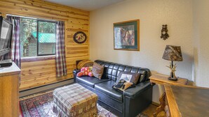 A cozy cabin interior with a wooden wall, a black leather sofa, a plaid rug, and a television on a wooden stand. - Island Park Fishing Cabin - Walk to Henrys Fork! (Island Park)