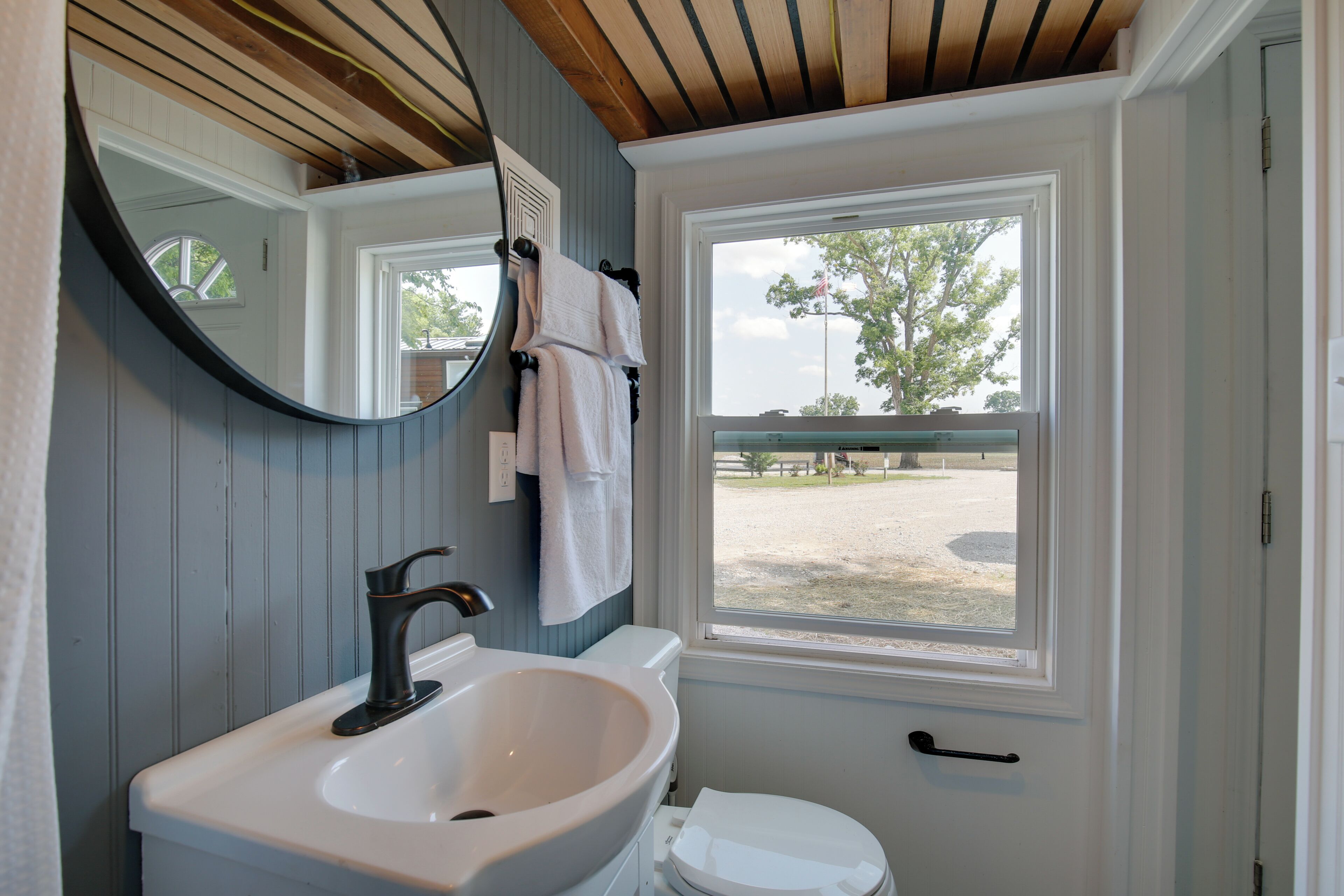 Ein Badezimmer mit einem weißen Waschbecken, einem Spiegel, einer weißen Toilette und einem Fenster mit Blick auf einen eingezäunten Bereich.