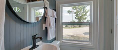 Ein Badezimmer mit einem weißen Waschbecken, einem Spiegel, einer weißen Toilette und einem Fenster mit Blick auf einen eingezäunten Bereich.
