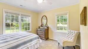 Un dormitorio con cama, cómoda, silla y ventanas amplias.