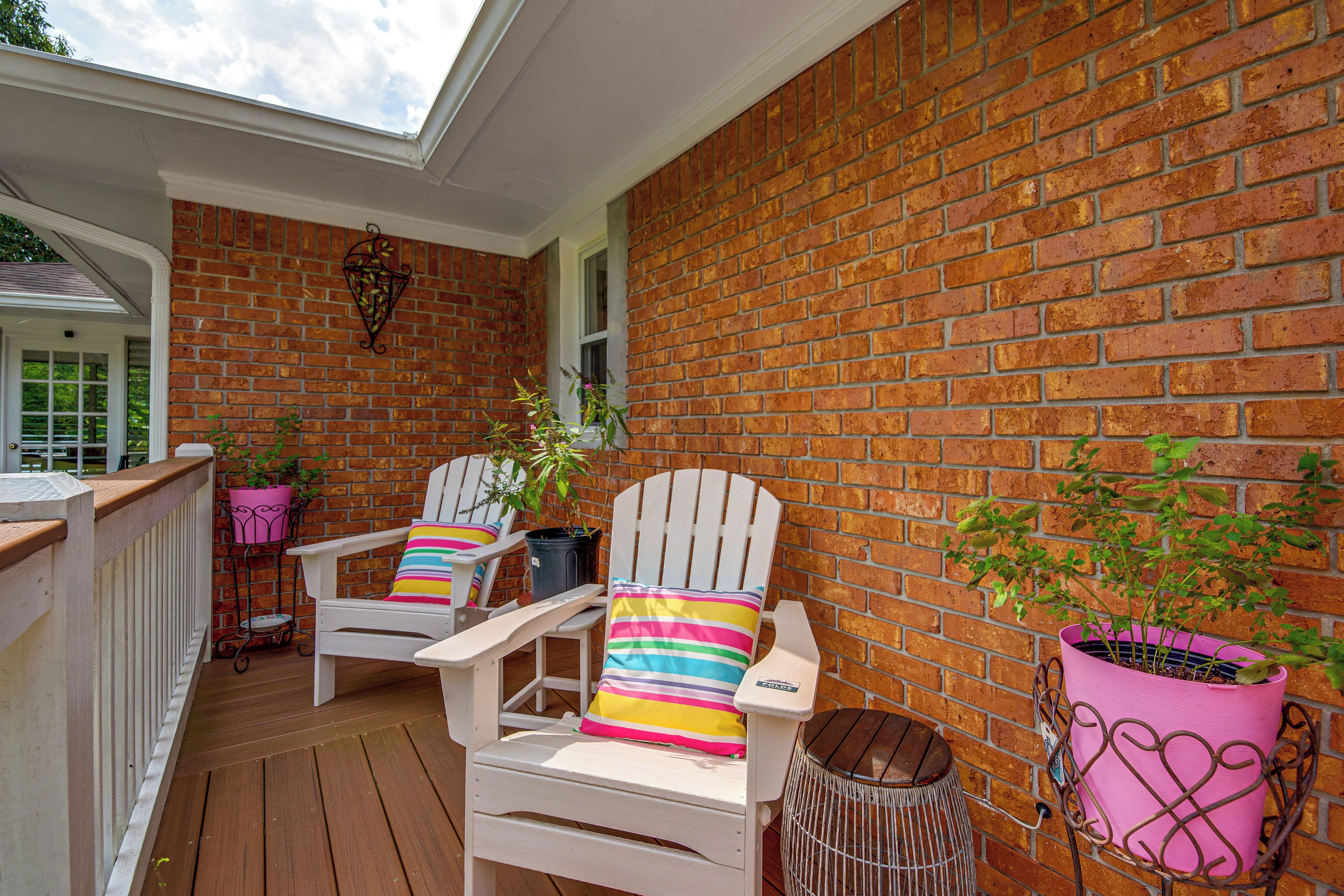 A covered porch with two Adirondack chairs, a brick wall, and potted plants.