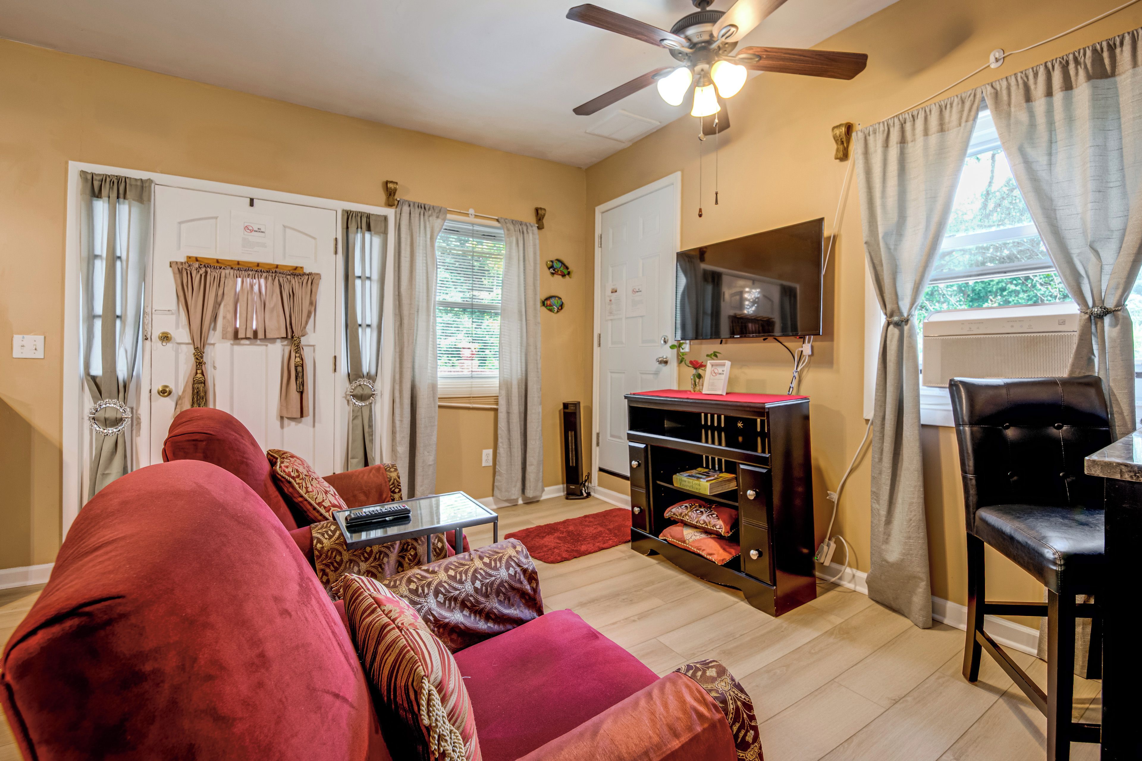 A living room with a red sofa, a small table, a ceiling fan, and a TV on the wall.