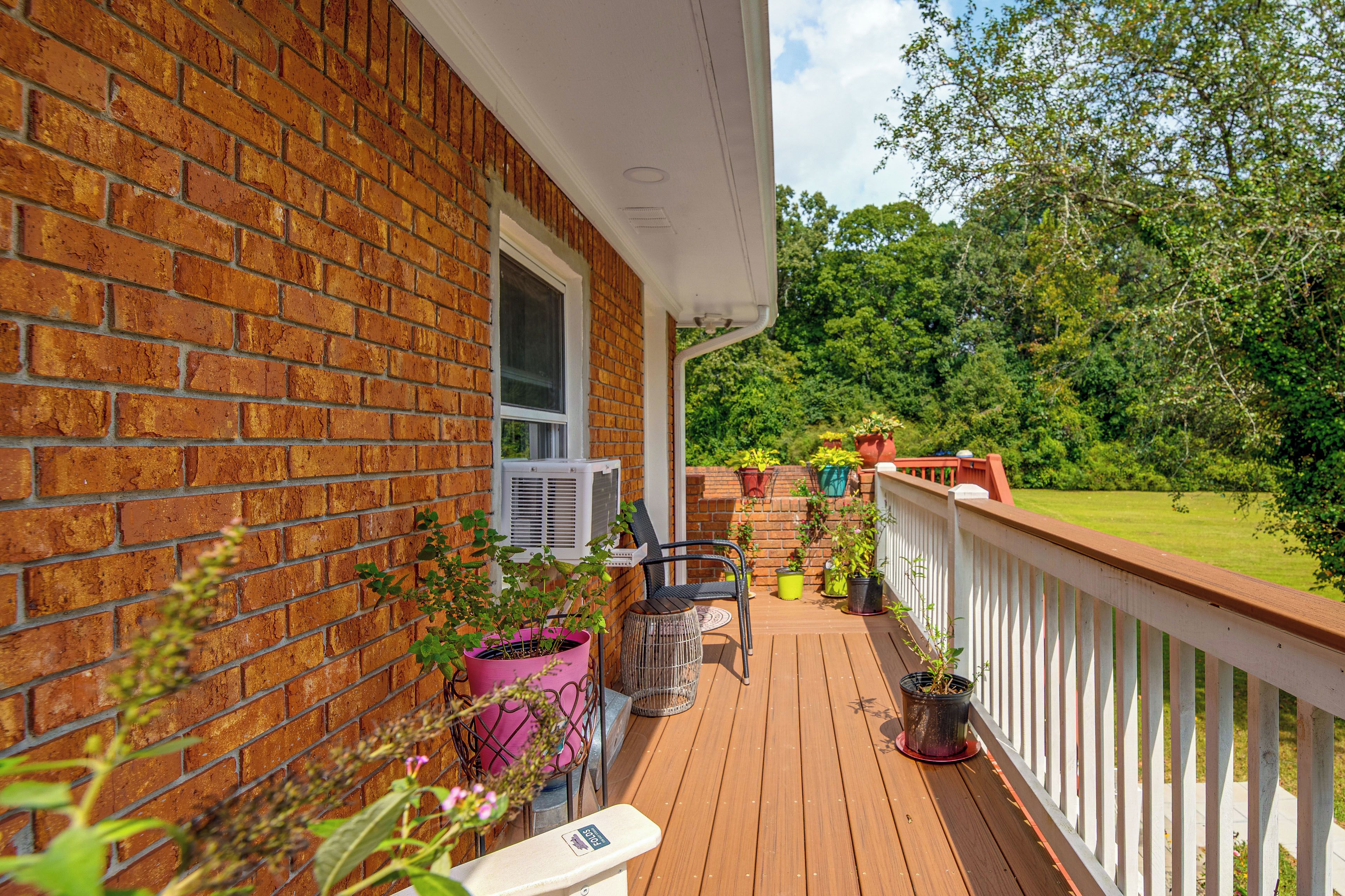 Une terrasse en bois avec une balustrade blanche, des plantes en pot et une fenêtre équipée d’un appareil de climatisation.