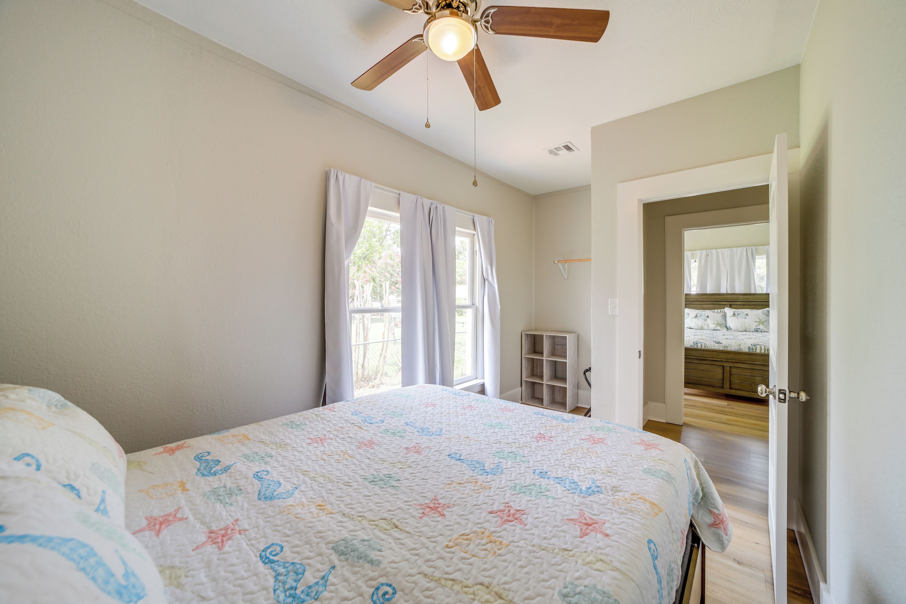 A bedroom with a bed, a ceiling fan, a window with curtains, and a door leading to another room.
