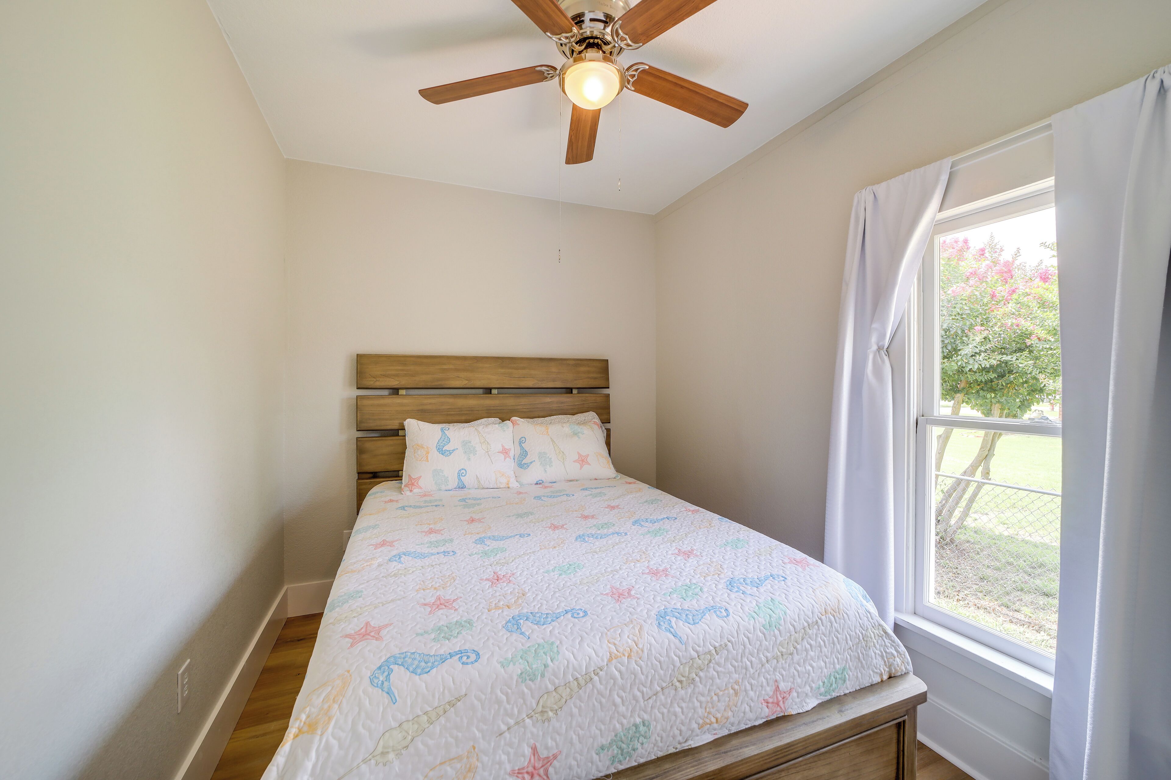 Un dormitorio con cabecera de madera, cama con colcha de estampado de estrellas, ventilador de techo y ventana con cortinas.