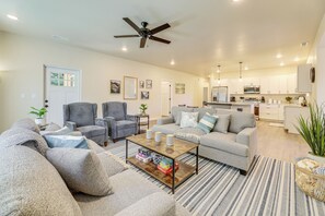 Una sala de estar moderna con sofá, sillones y una mesa de centro. Se ve una cocina al fondo con armarios blancos y un microondas.