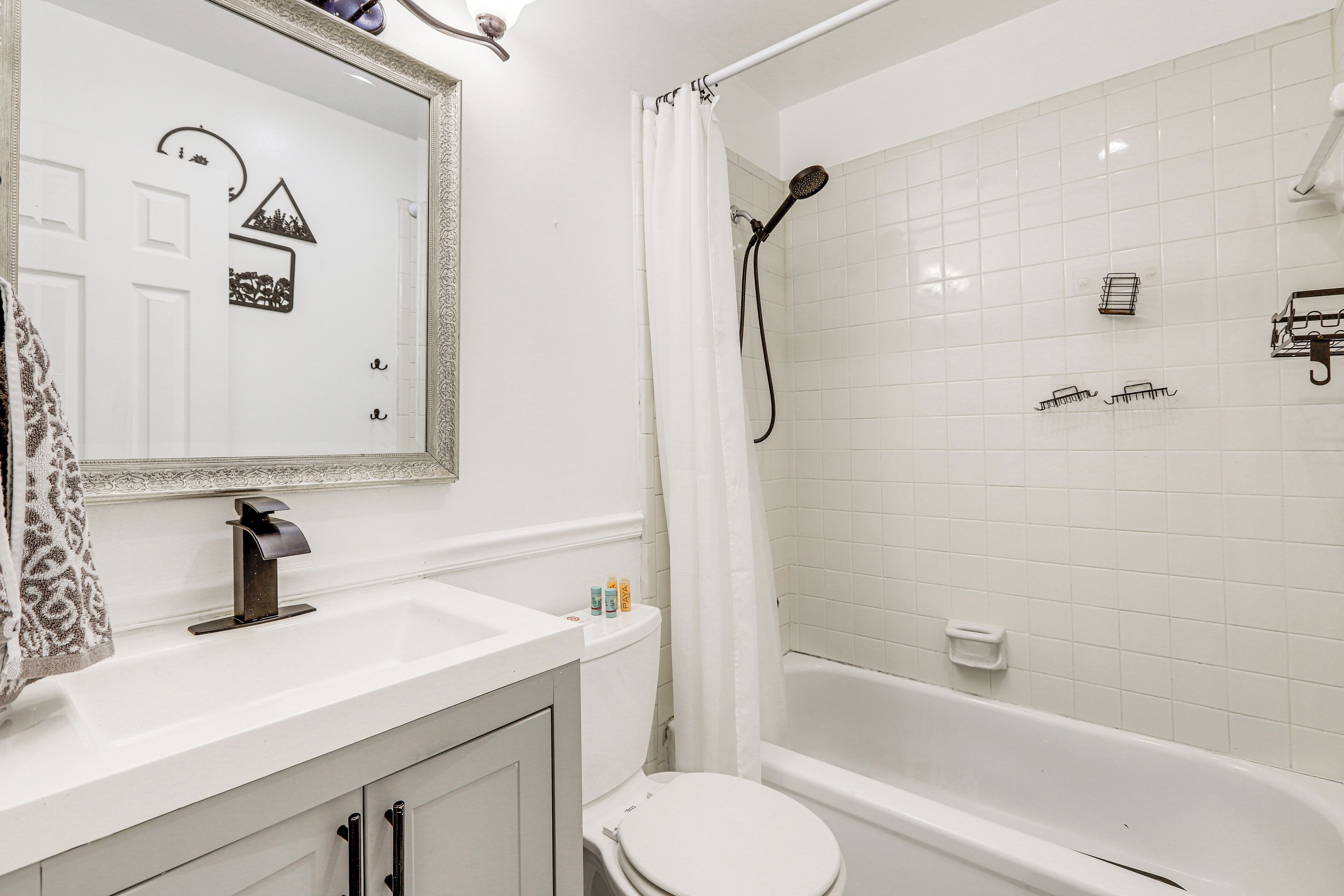 Ein Badezimmer mit einem weißen Waschbecken, einer Toilette und einer Badewanne. Ein Duschvorhang, ein Spiegel mit dekorativem Rahmen und ein Handtuch sind an der Wand angebracht.