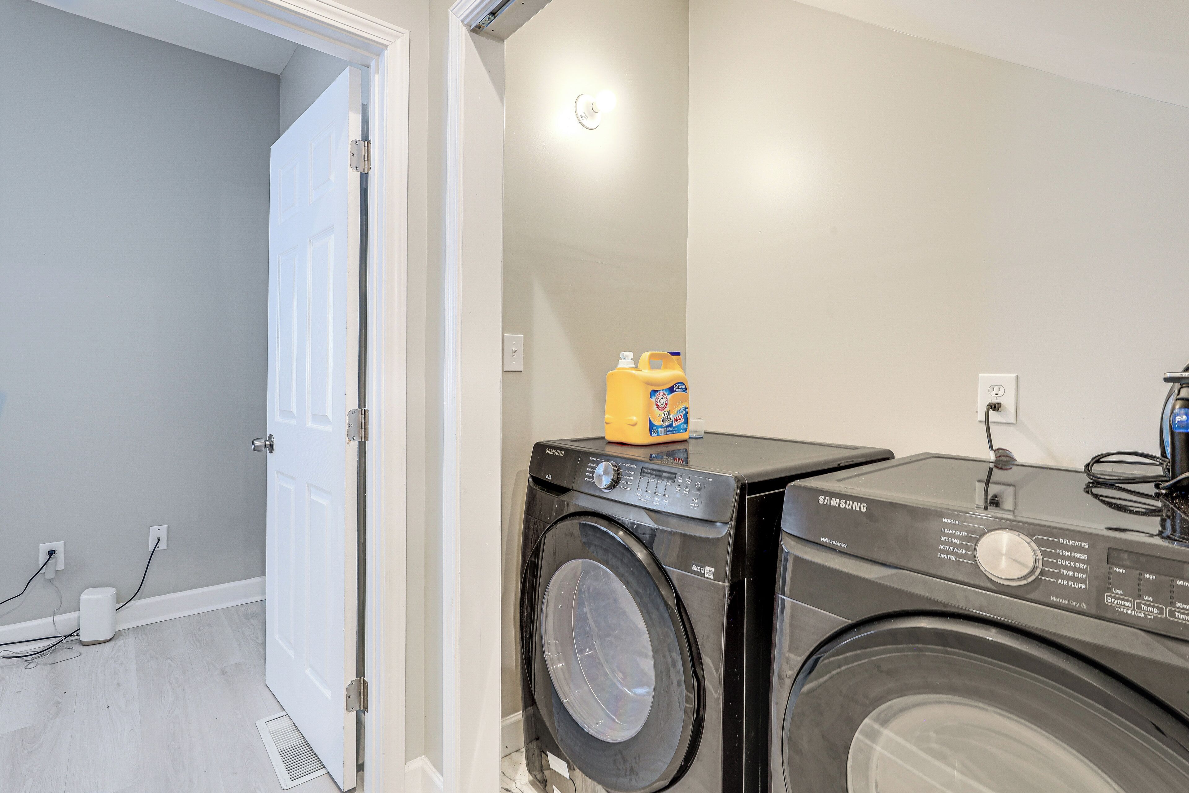 A laundry room with two front-loading washing machines, a bottle of detergent, and a power outlet on the wall.