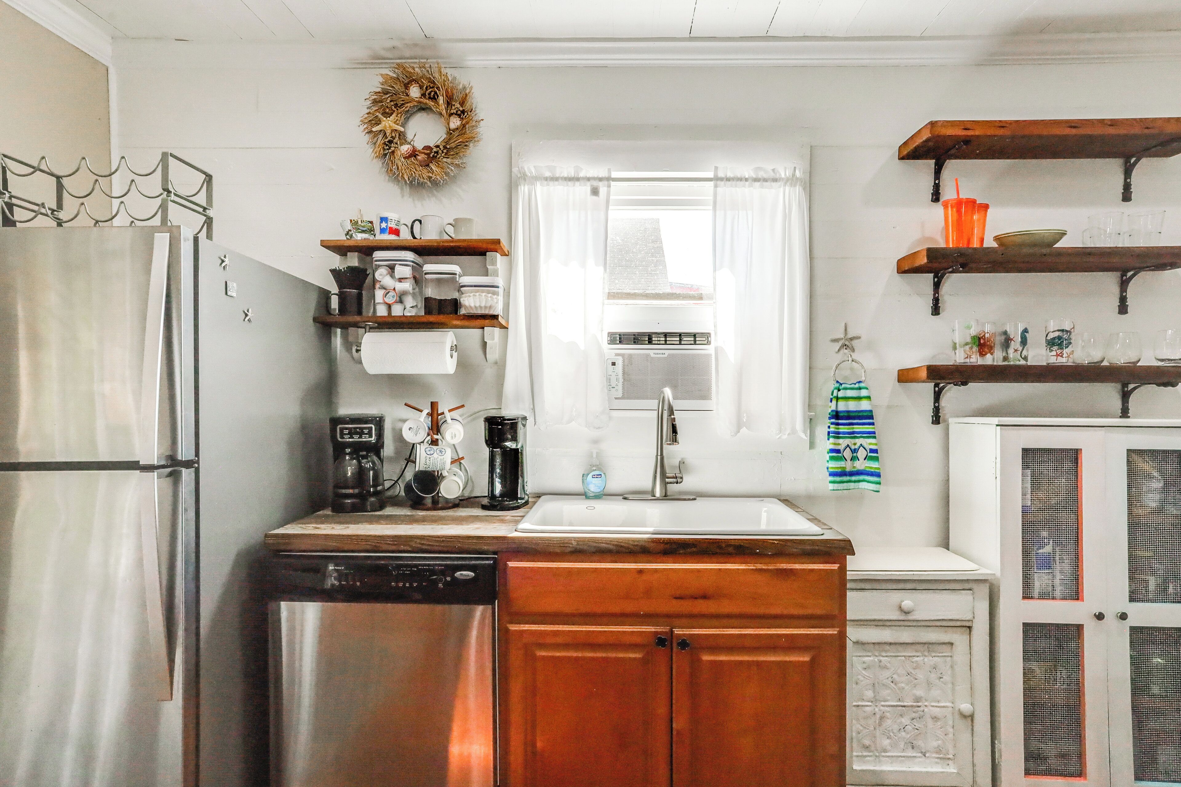 A kitchen with a stainless steel refrigerator, a dishwasher, wooden cabinets, a sink, and a window with a curtain. There are shelves above the sink and a decorative wreath on the wall.