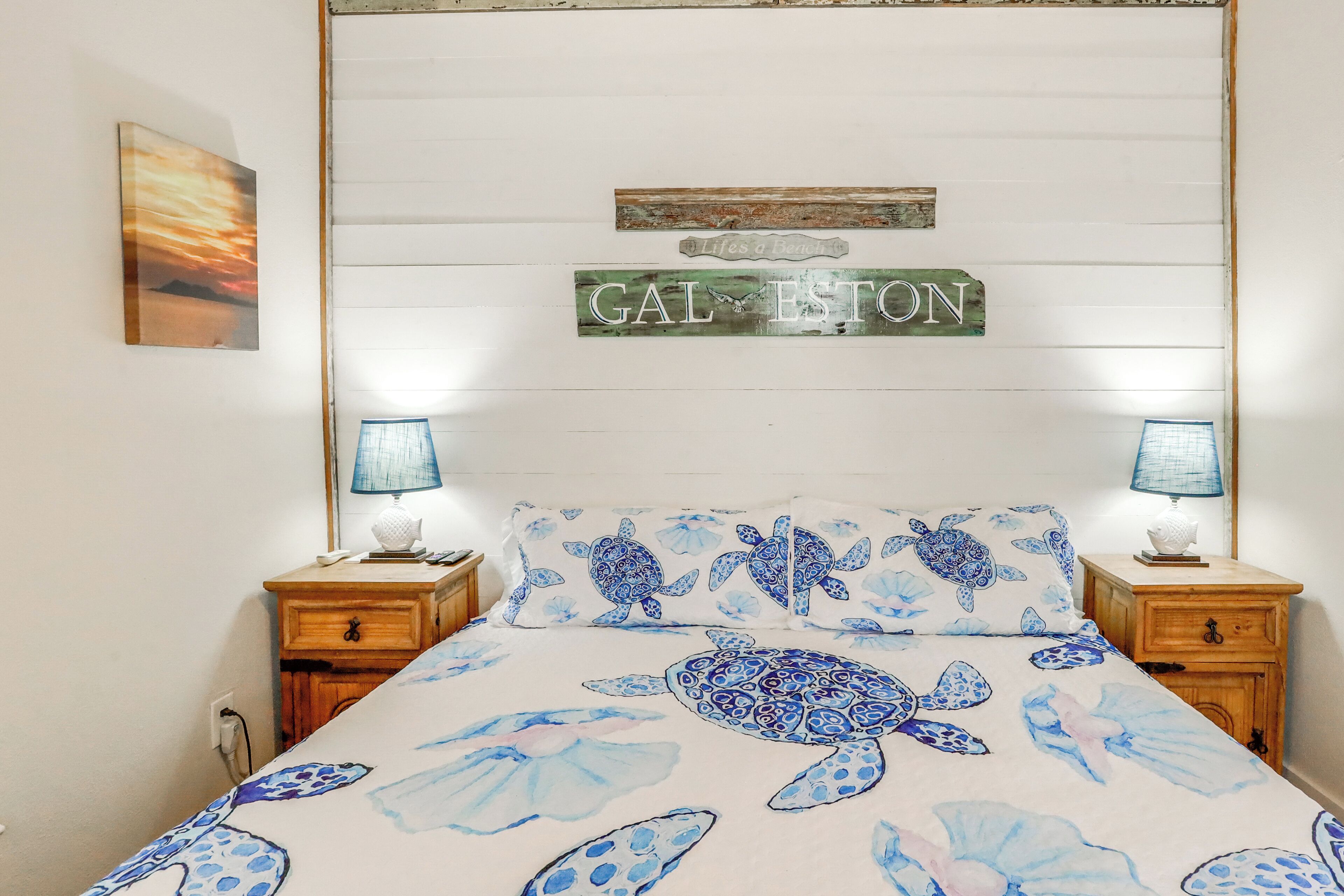 A bedroom with a bed featuring a sea turtle print, two bedside tables with lamps, a wall-mounted sign reading "GAL ESTON," and a framed picture above the bed.