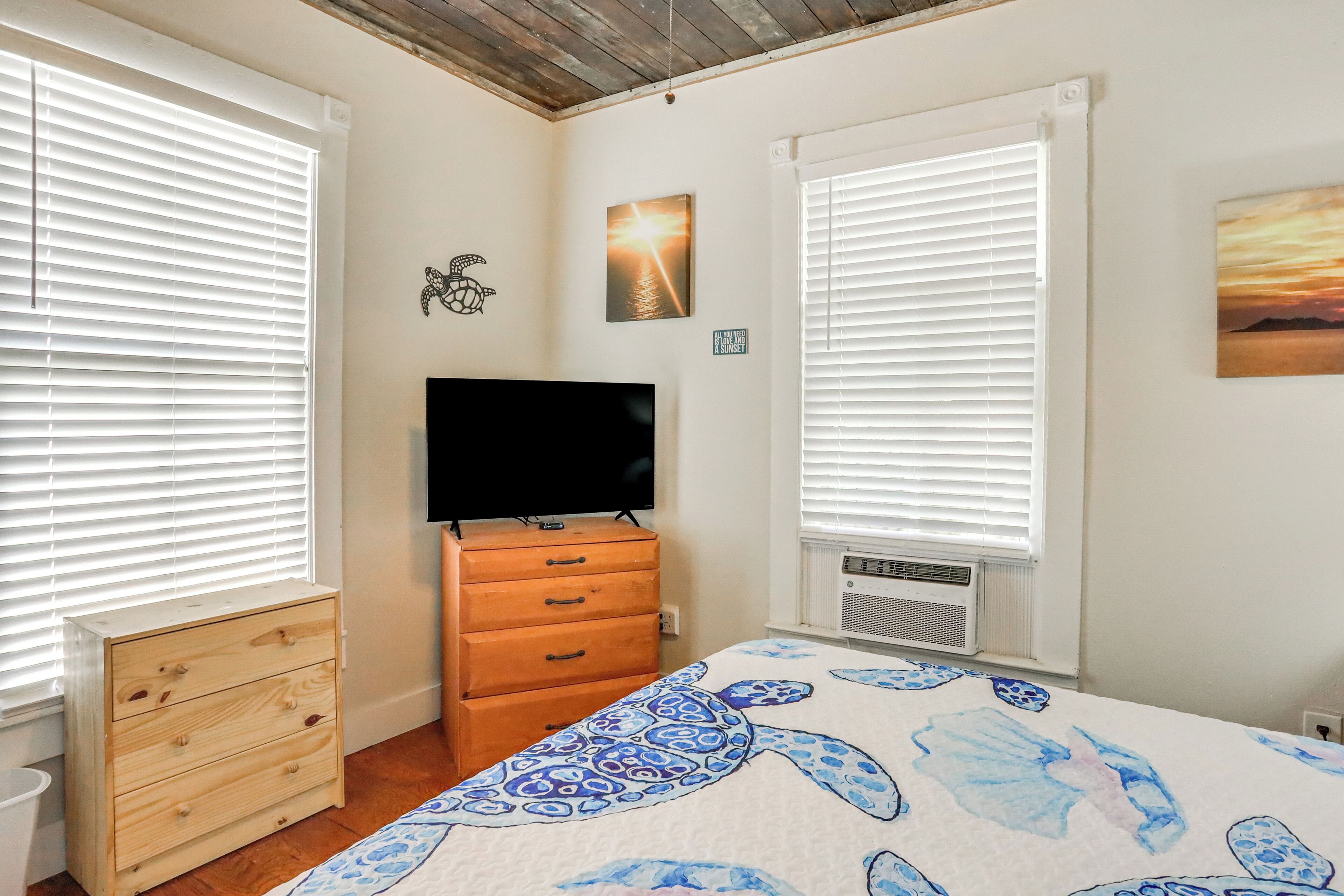 A bedroom with a bed, dresser, TV, and two windows with blinds.