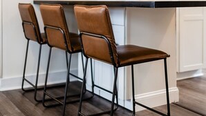 Una cocina moderna con piso de madera oscura, encimera blanca y tres sillas de cuero marrón con estructura de metal negro.