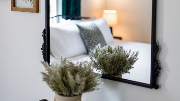 Un espejo con marco decorativo, un jarrón con plantas secas, y una cama con ropa blanca y una cortina verde en el reflejo.