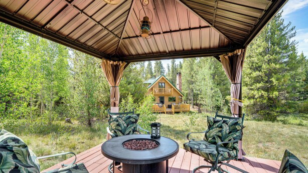 A covered outdoor seating area with a fire pit, surrounded by chairs with leaf-patterned cushions, overlooking a forested area and a cabin. - 24str-00023 Fairplay Cabin w/ Hot Tub on 4 Acres! (Fairplay)