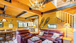 A cozy living room with a leather sofa, wooden dining table, and a staircase. - 24str-00023 Fairplay Cabin w/ Hot Tub on 4 Acres! (Fairplay)