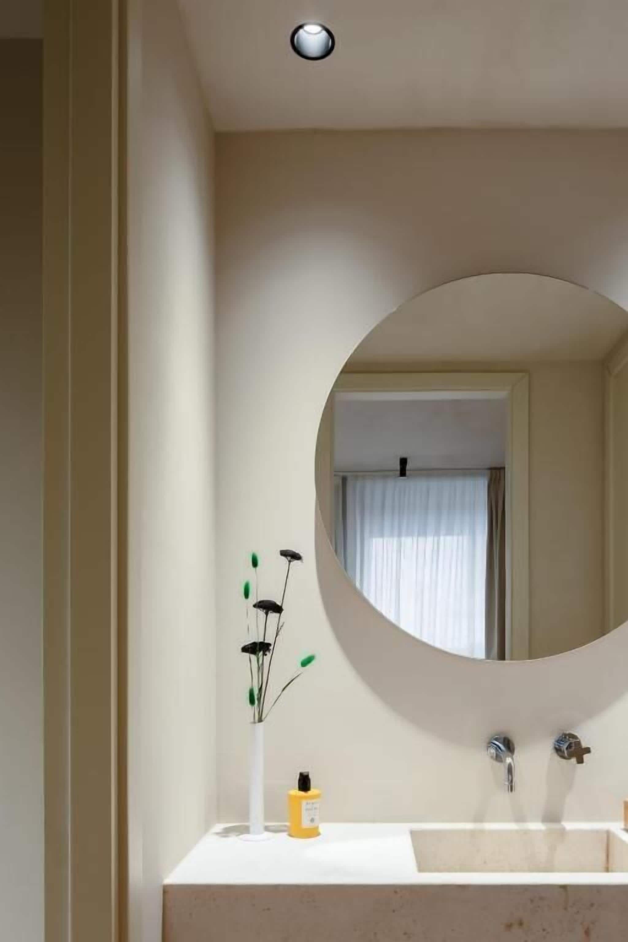 A bathroom with a round mirror, a white sink, and a vase with flowers.