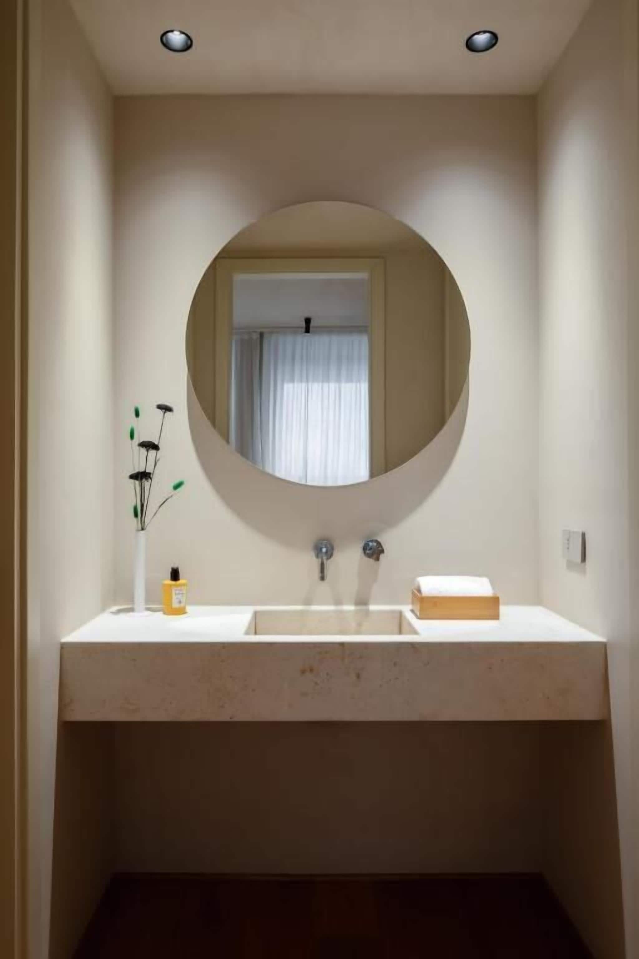 A bathroom with a round mirror, a white sink, and a window with curtains.