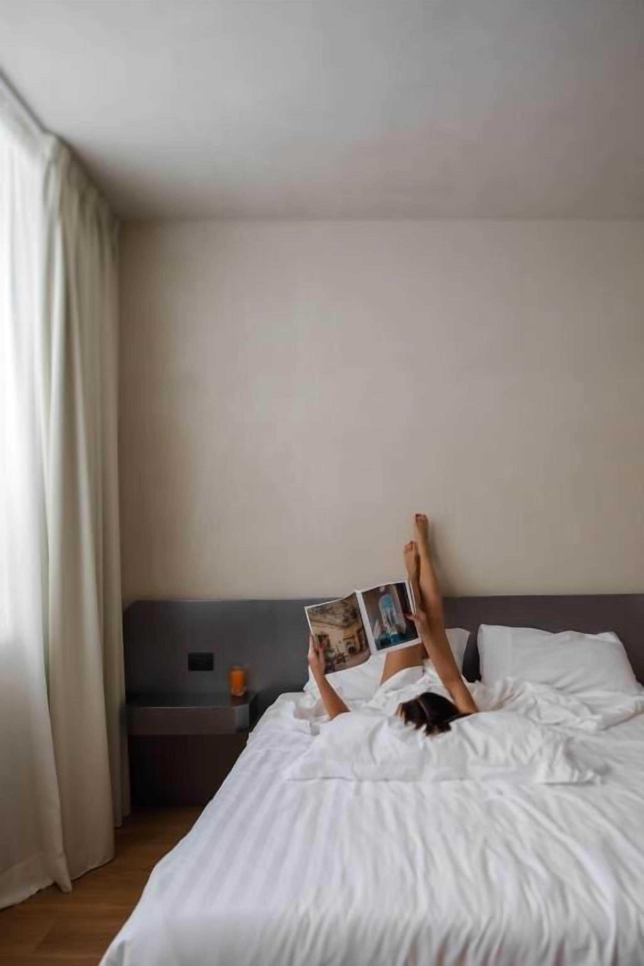 Una persona acostada en la cama leyendo un libro en una habitación con una mesita de noche y una ventana con cortinas.