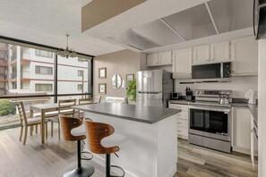 Una cocina moderna con una isla central, electrodomésticos de acero inoxidable y vista a un edificio residencial.