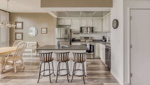 Una cocina moderna con armarios blancos, una isla central con taburetes, electrodomésticos de acero inoxidable y un comedor con mesa y sillas de madera.