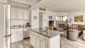 Una cocina moderna con armarios blancos, un refrigerador de acero inoxidable y una isla central. Hay una sala de estar con un sofá de cuero marrón y una chimenea.