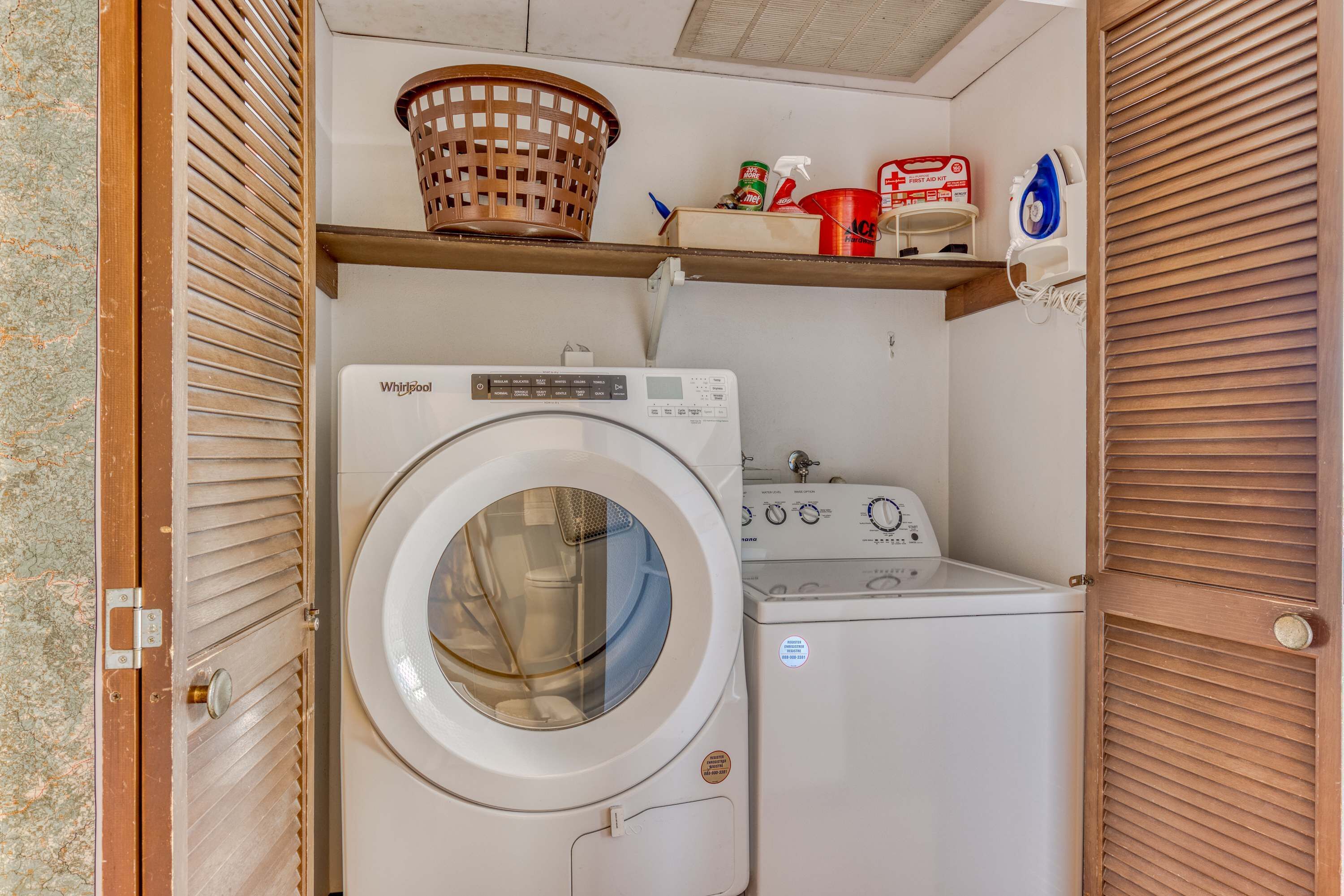 A compact laundry room with a front-loading washer and dryer, a wooden shelf with cleaning supplies, and a basket above the dryer.