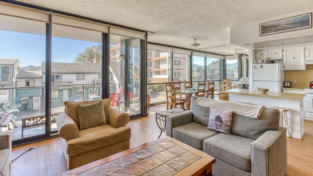 A spacious living area with a large sliding glass door leading to a balcony, a kitchen area, and a seating area with two sofas and a coffee table. - Sand & Sea (116) Columbia - Cozy Beach Getaway (Seaside)