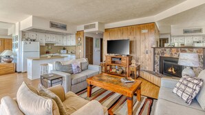 A spacious living area with a sofa, coffee table, and fireplace. A kitchen with white cabinets and a refrigerator is visible in the background. - Sand & Sea (116) Columbia - Cozy Beach Getaway (Seaside)