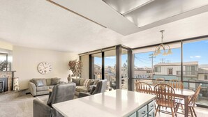 A spacious living area with a large window, a fireplace, and a dining area with wooden chairs and a table. - Sand & Sea (118) Shore Thing - Oceanside Charm (Seaside)