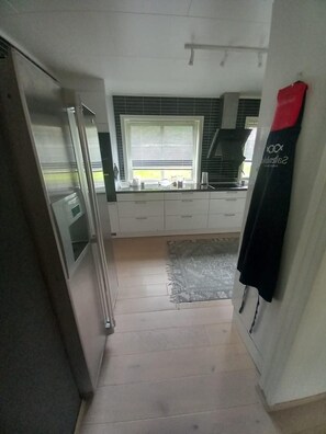 A narrow kitchen with a refrigerator, a window with blinds, a countertop with kitchen appliances, and a rug on the floor.