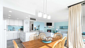 A modern kitchen with white cabinets, a wooden dining table with chairs, and a view of the ocean through large windows. - Sand & Sea (214) Turtle Cove - Condo on the Prom w Pool (Seaside)