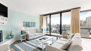A spacious living room with a large window, a sofa set, a coffee table, and a fireplace. - Sand & Sea (214) Turtle Cove - Condo on the Prom w Pool (Seaside)