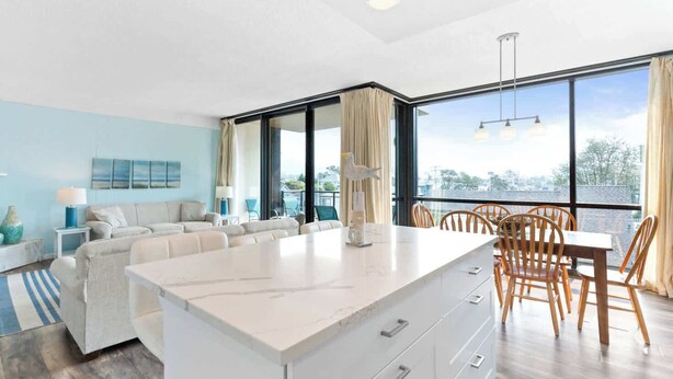 A modern living room with a large window, a sofa set, a dining table with chairs, and a white cabinet. - Sand & Sea (214) Turtle Cove - Condo on the Prom w Pool (Seaside)