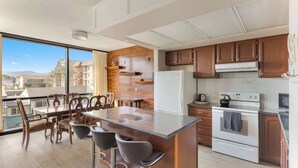 Eine moderne Küche mit Holzschränken, einer großen Kochinsel mit Barhocker, einem Kühlschrank, einem Backofen und Blick durch große Fenster.