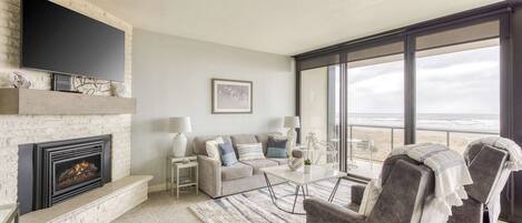A modern living room with a fireplace, a flat-screen TV, a sofa, two armchairs, a coffee table, and large windows with a view of the beach.