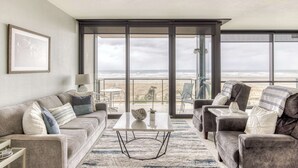 A modern living room with a sofa, armchairs, and a coffee table. There is a large window with a view of the beach. - Sand & Sea (306) Sojourn - Oceanfront luxury (Seaside)
