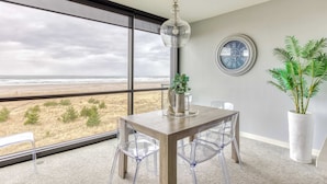 A modern dining area with a view of the beach, a large window, and a potted plant. - Sand & Sea (306) Sojourn - Oceanfront luxury (Seaside)