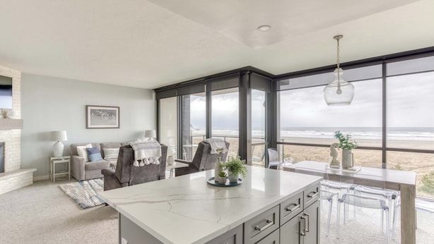 A modern kitchen with a large island, a dining area with clear chairs, and a view of the beach. - Sand & Sea (306) Sojourn - Oceanfront luxury (Seaside)