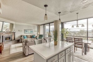 A modern living room with a fireplace, a sofa set, a dining area with a table and chairs, and large windows offering a view. - Sand & Sea (312) Coastal Range - Pet Friendly (Seaside)