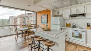A modern kitchen with white cabinets, a central island with stools, a large window, and a view of neighboring buildings.